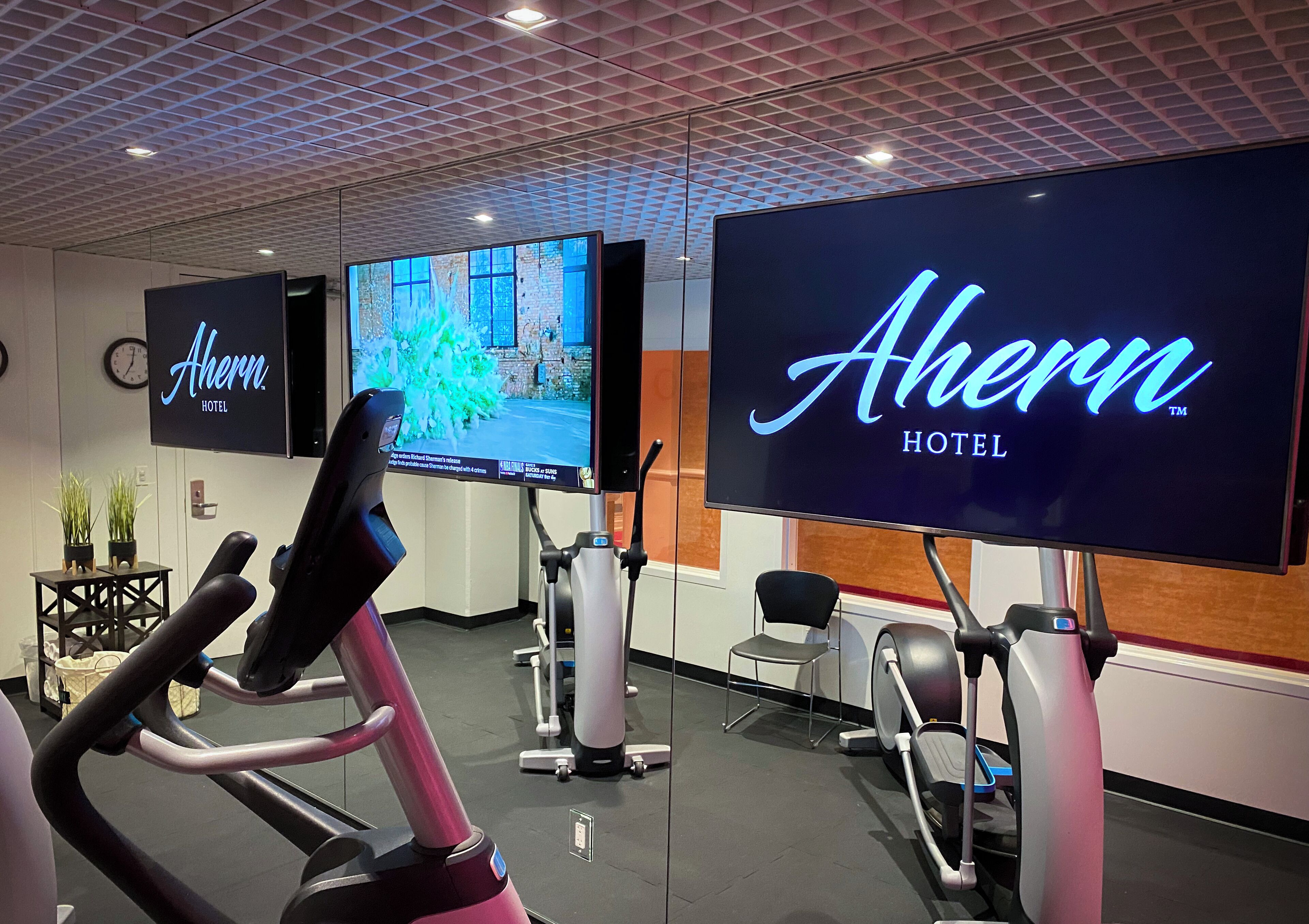 Et fitnessområde med træningsudstyr, herunder et løbebånd og en crosstrainer, samt to store skærme, der viser hotellets navn.