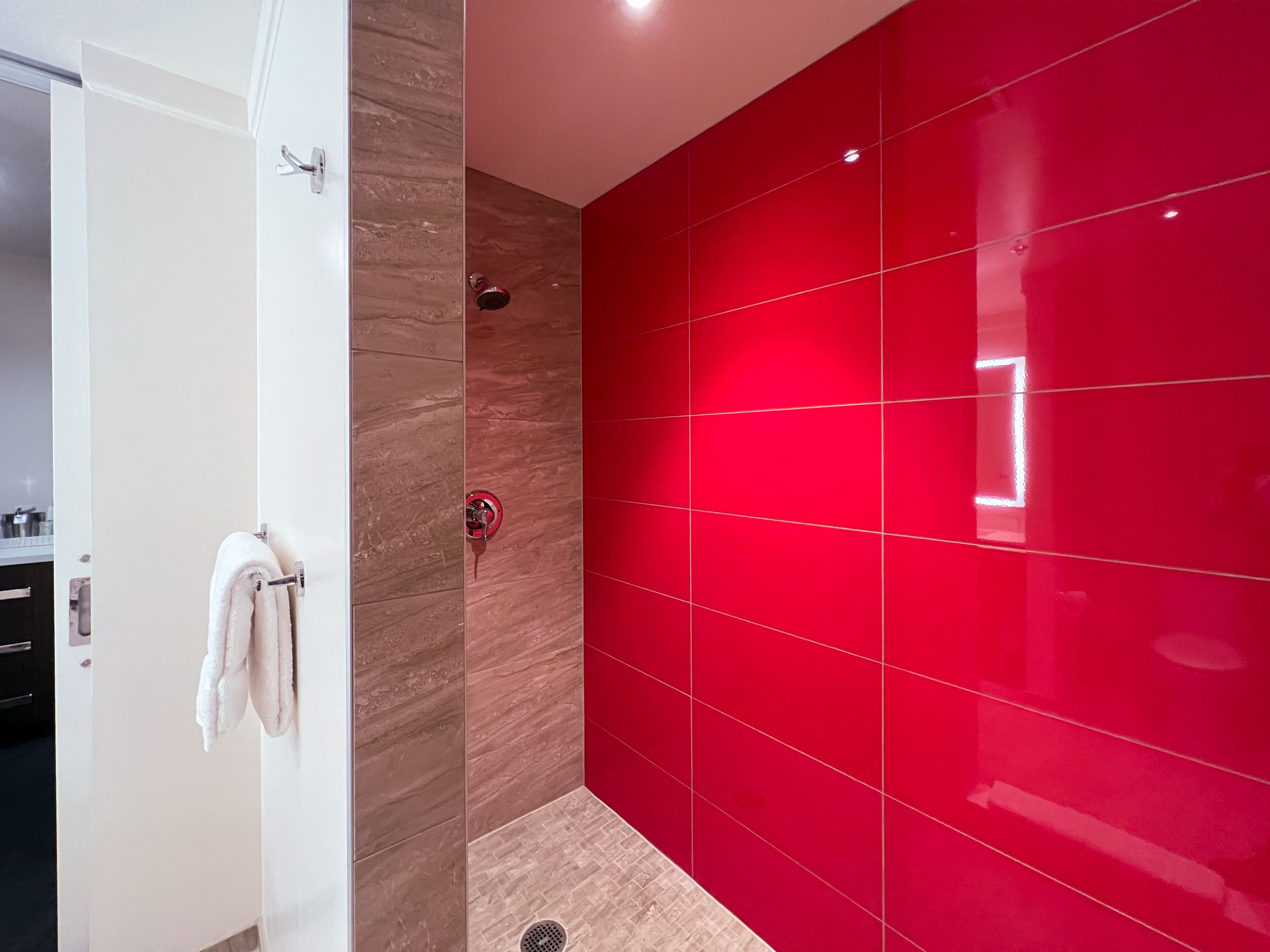 Un baño moderno con una puerta blanca, un perchero, una ducha con pared roja y piso de baldosas.
