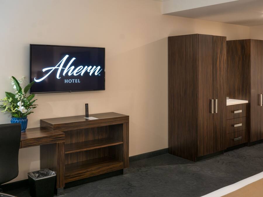 Un espace de réception d’hôtel comprenant un téléviseur à écran plat affichant le nom de l’hôtel, un bureau en bois avec une poubelle et une grande armoire avec des tiroirs.