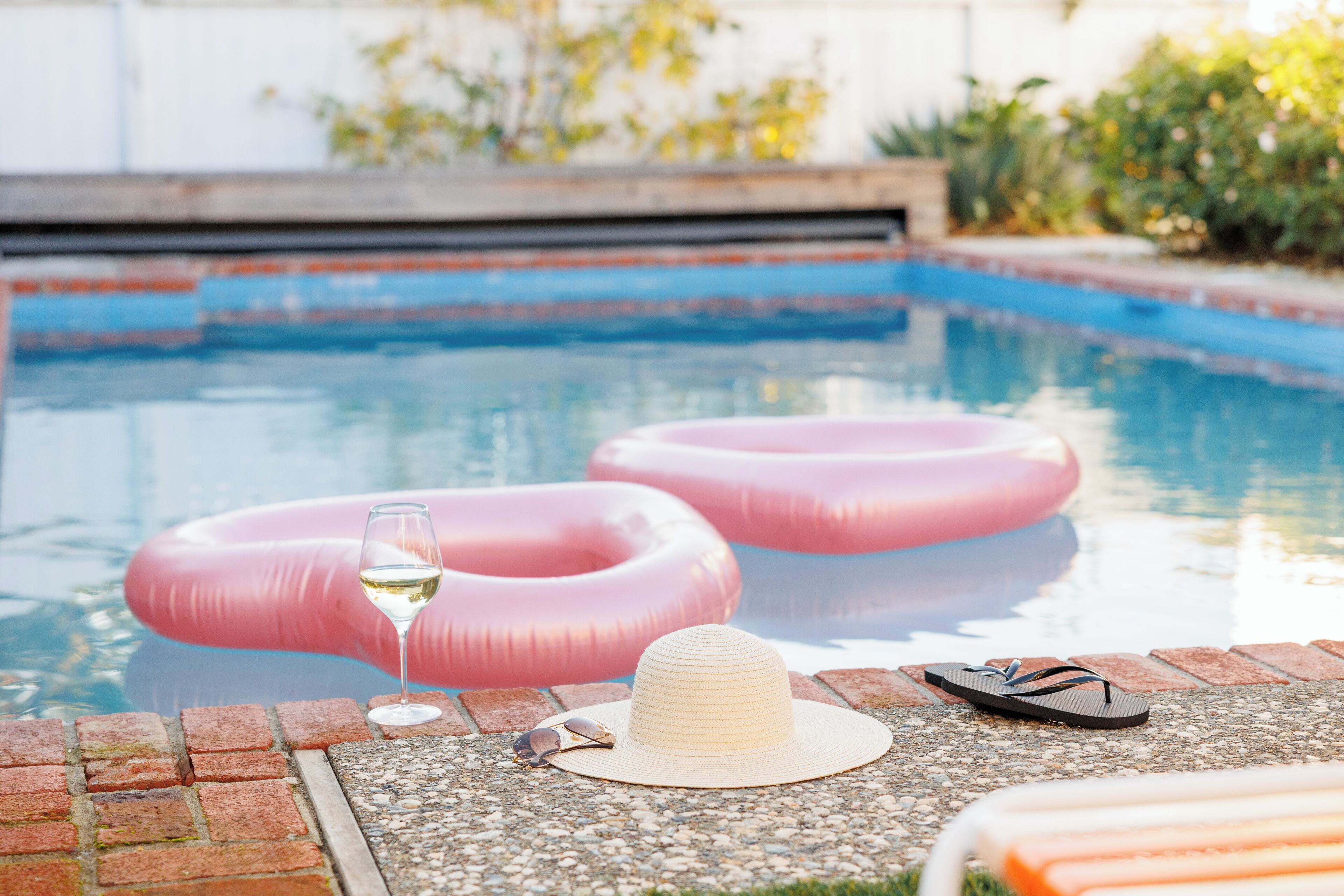A pool area with two pink inflatable rings, a glass of white wine, a straw hat, and a pair of flip-flops on the edge.