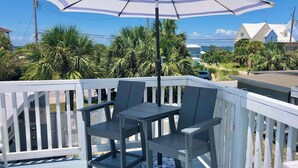 Un balcon avec une table et des chaises, un parasol blanc, et une vue sur des palmiers et des maisons.