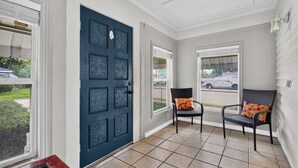 Un porche cubierto con una puerta azul oscuro, dos sillas de mimbre con cojines, y vista a un estacionamiento a través de las ventanas.