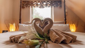 Ein ordentlich bezogenes Bett mit Handtüchern, die zu einem Herz geformt sind, einer weißen Blume und einem Fenster mit Blick auf Bäume.