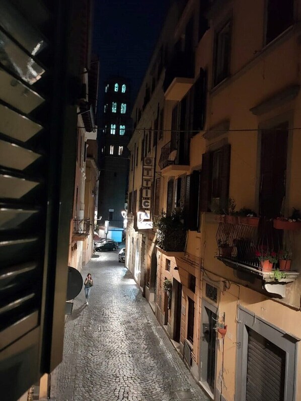 Uma rua de paralelepípedos estreita, ladeada por edifícios com varandas e toldos, conduzindo a um prédio com sinalização iluminada.