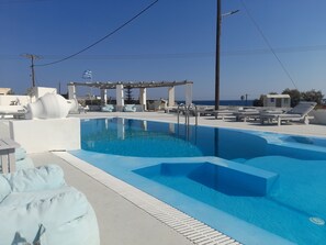 A swimming pool with lounge chairs and a pergola, surrounded by a white pool deck and a view of the sea. - Sea paradise villas (Santorini)