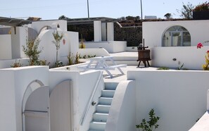 A white-walled building with a staircase leading to a covered area, a wooden bench, and a small garden with plants. - Sea paradise villas (Santorini)