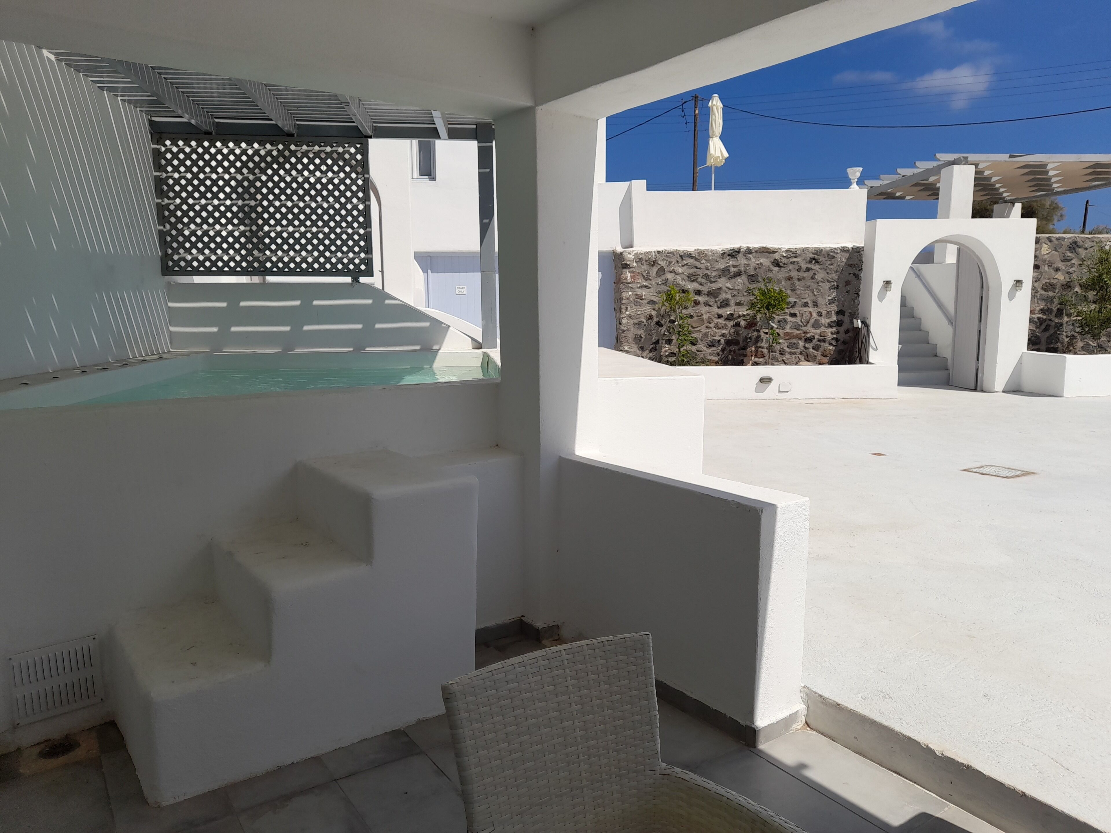Ein moderner Poolbereich im Freien mit einer Steinwand, einer weißen Pergola und einer Treppe, die zu einer überdachten Terrasse führt.