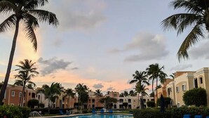A resort pool with palm trees and lounge chairs, surrounded by multi-story buildings. - Escape Beach Studio, Fairway Courts, Palmas del Mar (Humacao)