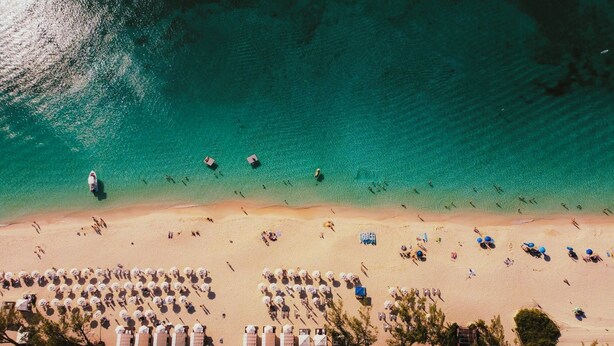 Eine Vogelperspektive zeigt einen Strand mit klarem türkisblauem Wasser, einem sandigen Ufer und zahlreichen Strandstühlen und Liegen.