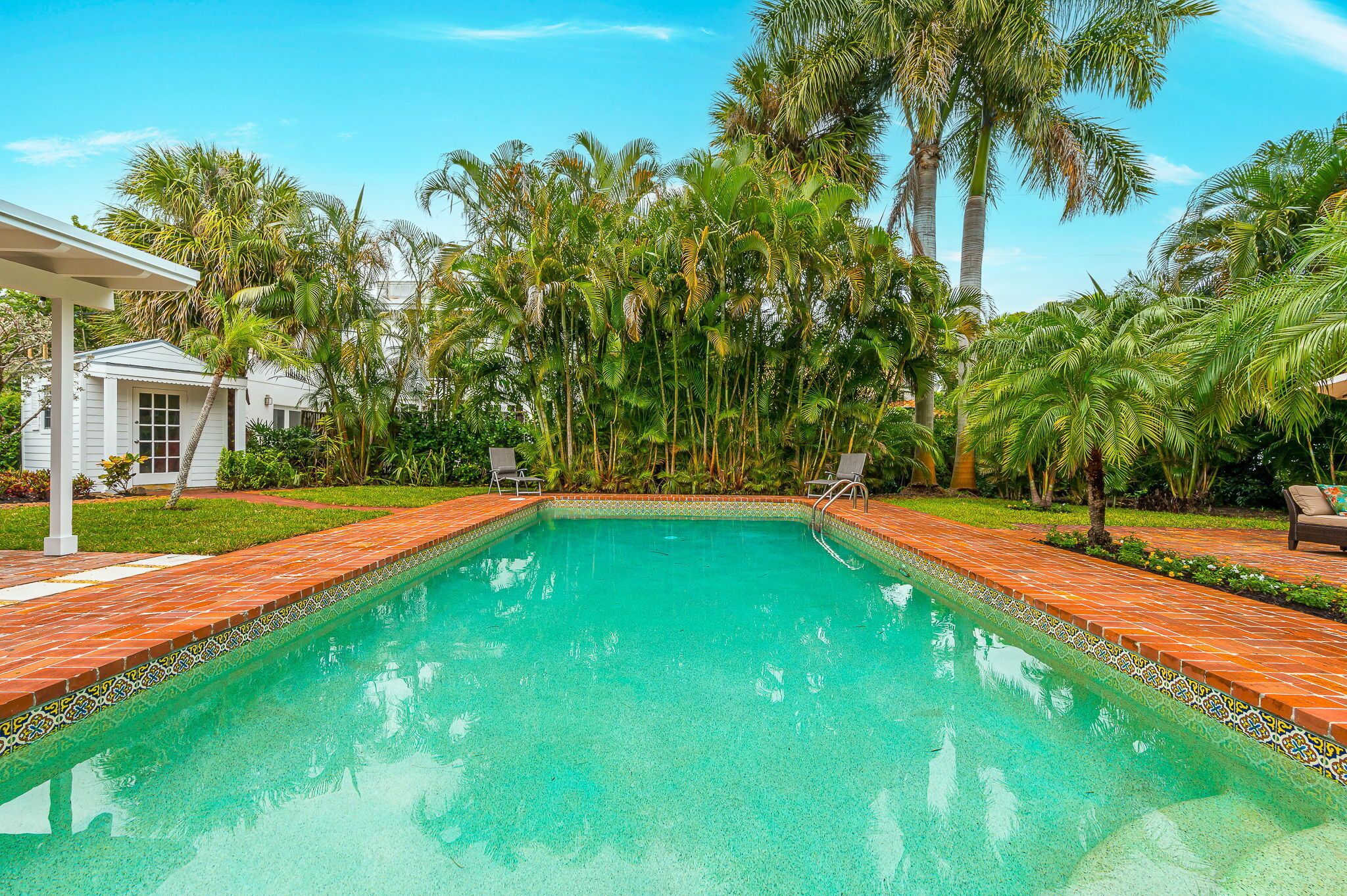 Une piscine rectangulaire bordée de carreaux, entourée de palmiers et d’un bâtiment blanc doté d’un patio couvert.