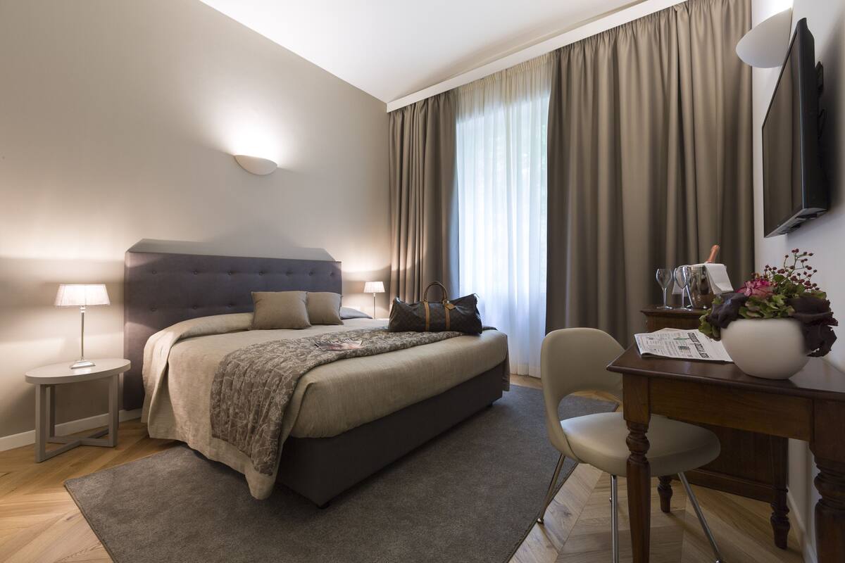 Quarto com cama, mesa de cabeceira, abajur, cadeira e escrivaninha com um vaso e papéis.
