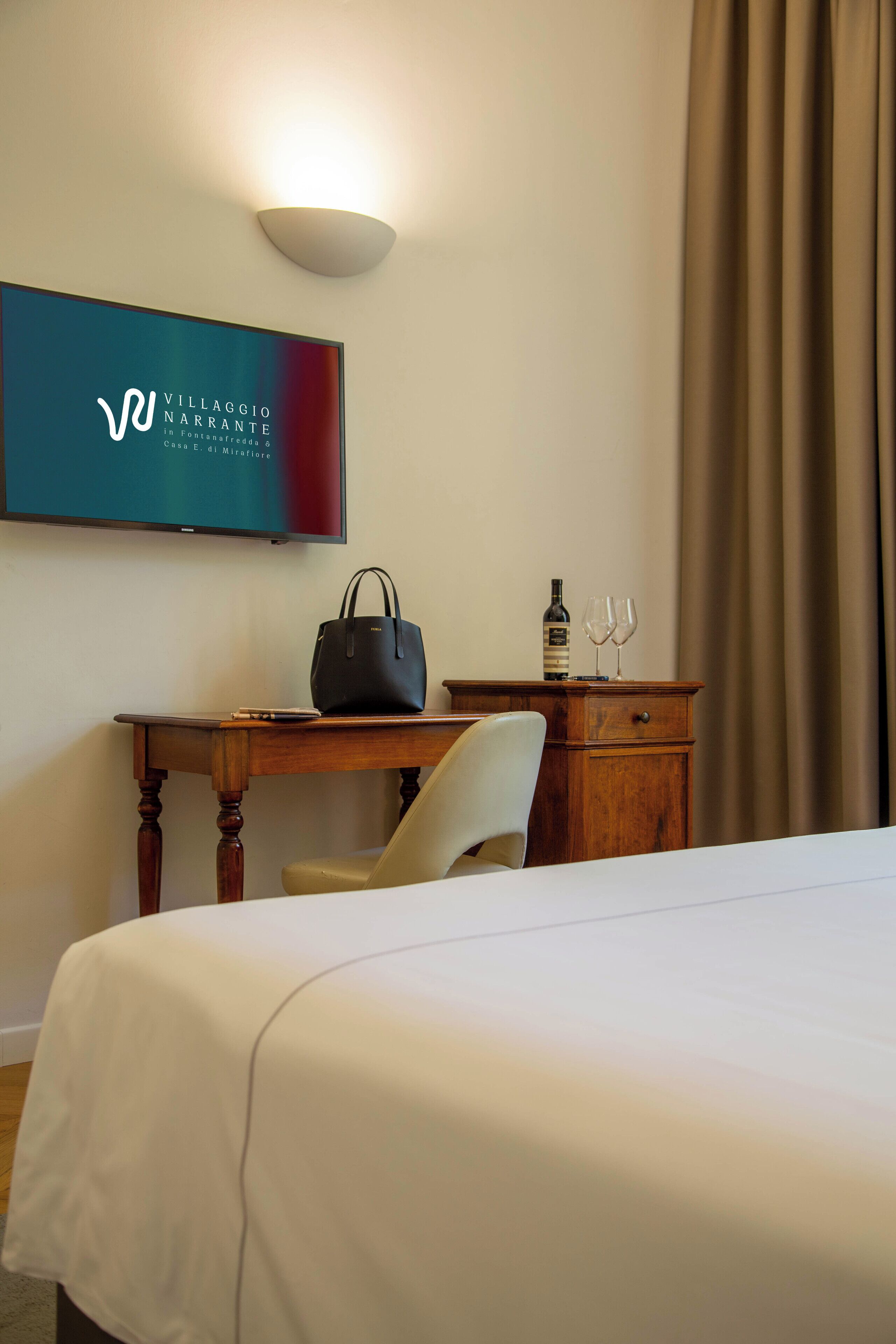 A hotel room with a bed, a desk with a chair, a television mounted on the wall, a bottle, and two wine glasses on the desk.