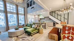 A spacious living area with a green sofa, a round coffee table, and a dining table with chairs. A kitchen is visible in the background, and there is a staircase leading to an upper level. - 4 Bedroom WOW w/heated floors, Ski out access to Snowbird and steam bath. (Alta)