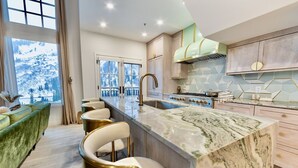 A modern kitchen with a marble island, bar stools, and a view of snow-covered mountains through large windows. - 4 Bedroom WOW w/heated floors, Ski out access to Snowbird and steam bath. (Alta)