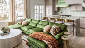 A modern living room with a green sectional sofa, a round coffee table, and a view of the kitchen area with a stove and bar stools. - 4 Bedroom WOW w/heated floors, Ski out access to Snowbird and steam bath. (Alta)