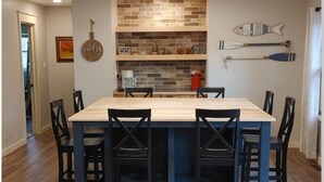 Un coin repas comprenant une table rectangulaire et six chaises, un mur recouvert de briques et des objets décoratifs disposés sur des étagères.