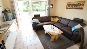 A living room with a brown leather sofa, a wooden coffee table, and a kitchen area with a sink and a television. - Vacation home "Sömmerwind 4" Norddeich, Riedeweg 12 (Norden)