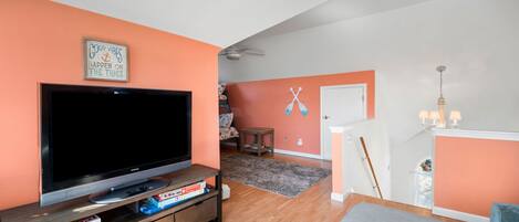 A living room with a flat-screen TV, a bookshelf with board games, a ceiling fan, and a door leading to another room.