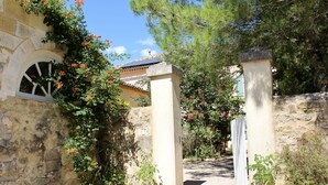 Ein gepflasterter Weg führt zu einem Gebäude mit Fenster und Tür, umgeben von Grünpflanzen und blühenden Blumen.
