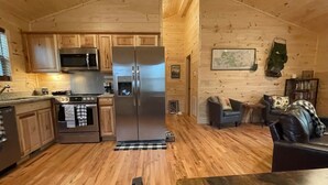 Ein Holzblockhaus-Interieur mit einer Küche, die Edelstahlgeräte, einen Essbereich mit einem Tisch und eine gemütliche Sitzecke mit einer Couch und einem Sessel beinhaltet.