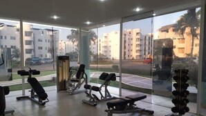 A gym with various exercise equipment, large windows, and a view of a residential area. - Rent New Apartment in Acapulco 5 min. from the airport beach and La Isla. (Acapulco de Juárez)