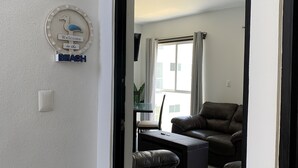 A modern living room with a leather sofa, ottoman, and a dining area with a table and chairs. A large window provides natural light, and the room features a clean, minimalist design. - Rent New Apartment in Acapulco 5 min. from the airport beach and La Isla. (Acapulco de Juárez)