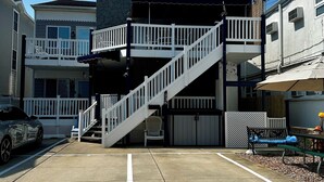 A multi-story building with a white staircase leading to a balcony, a garage, and a parking area with a car and a bench.