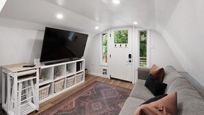 A modern living room with a flat-screen TV, white furniture, a patterned rug, and a view of greenery outside.