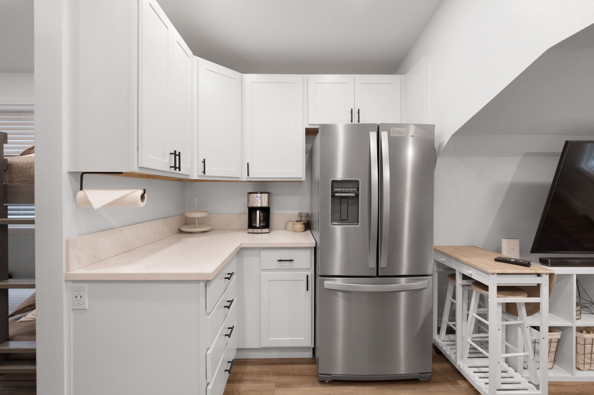 A modern kitchen with white cabinets, a stainless steel refrigerator, a coffee maker, and a wooden table.