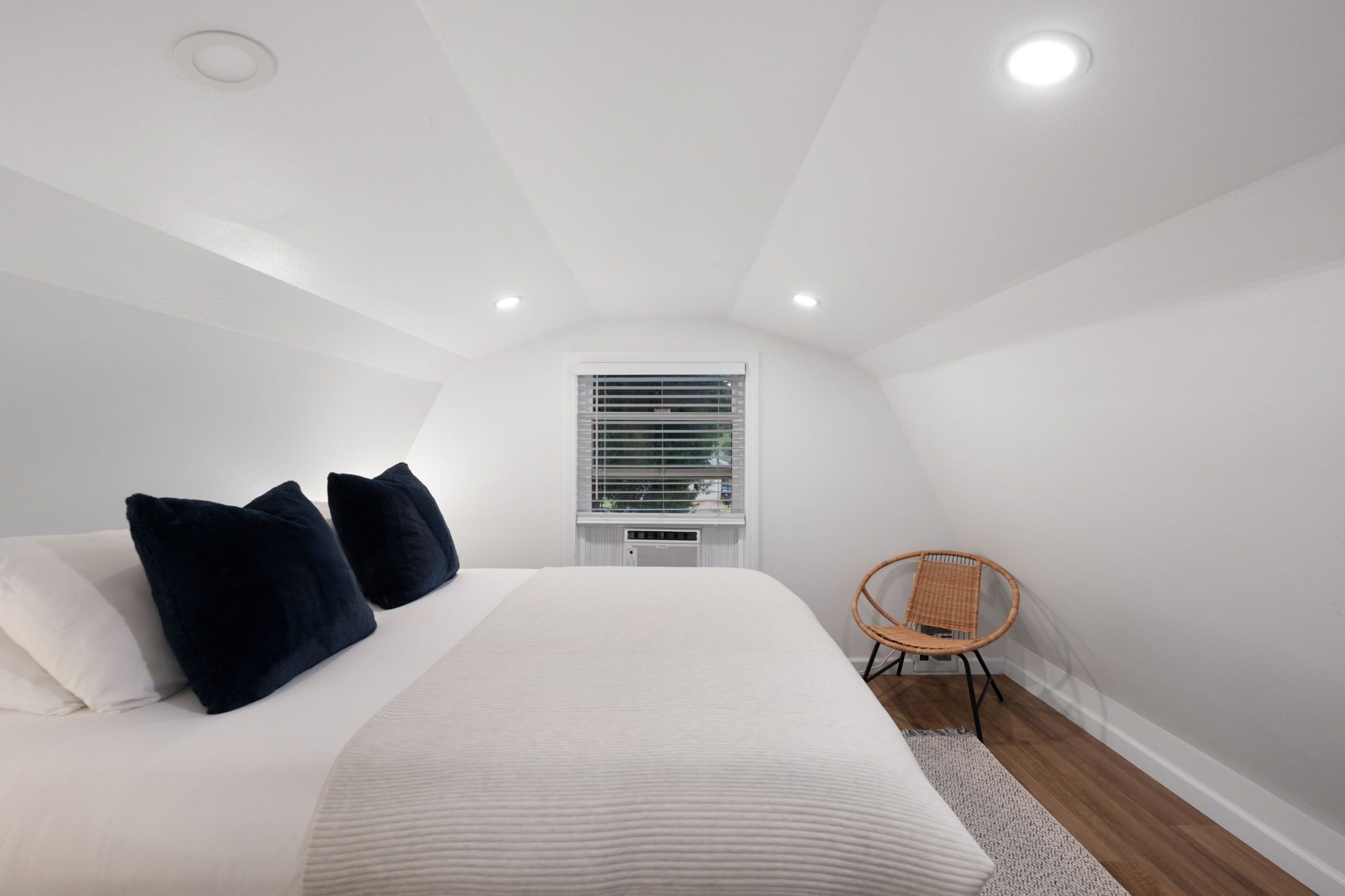 Un dormitorio con una cama grande, paredes blancas, una ventana con persianas, una silla y luces empotradas en el techo.