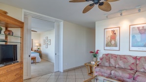 Uma sala de estar com sofá, mesa de centro com frutas, televisão e ventilador de teto.