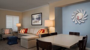 Een woonkamer met een beige bank, een blauwe poef, en een eettafel met vier stoelen. Er hangt een lijst met een afbeelding aan de muur en een lamp naast de bank.