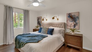 Un dormitorio con cabecera capitonada, una cama grande con ropa de cama blanca, dos mesitas de noche con lámparas y obras de arte en las paredes.