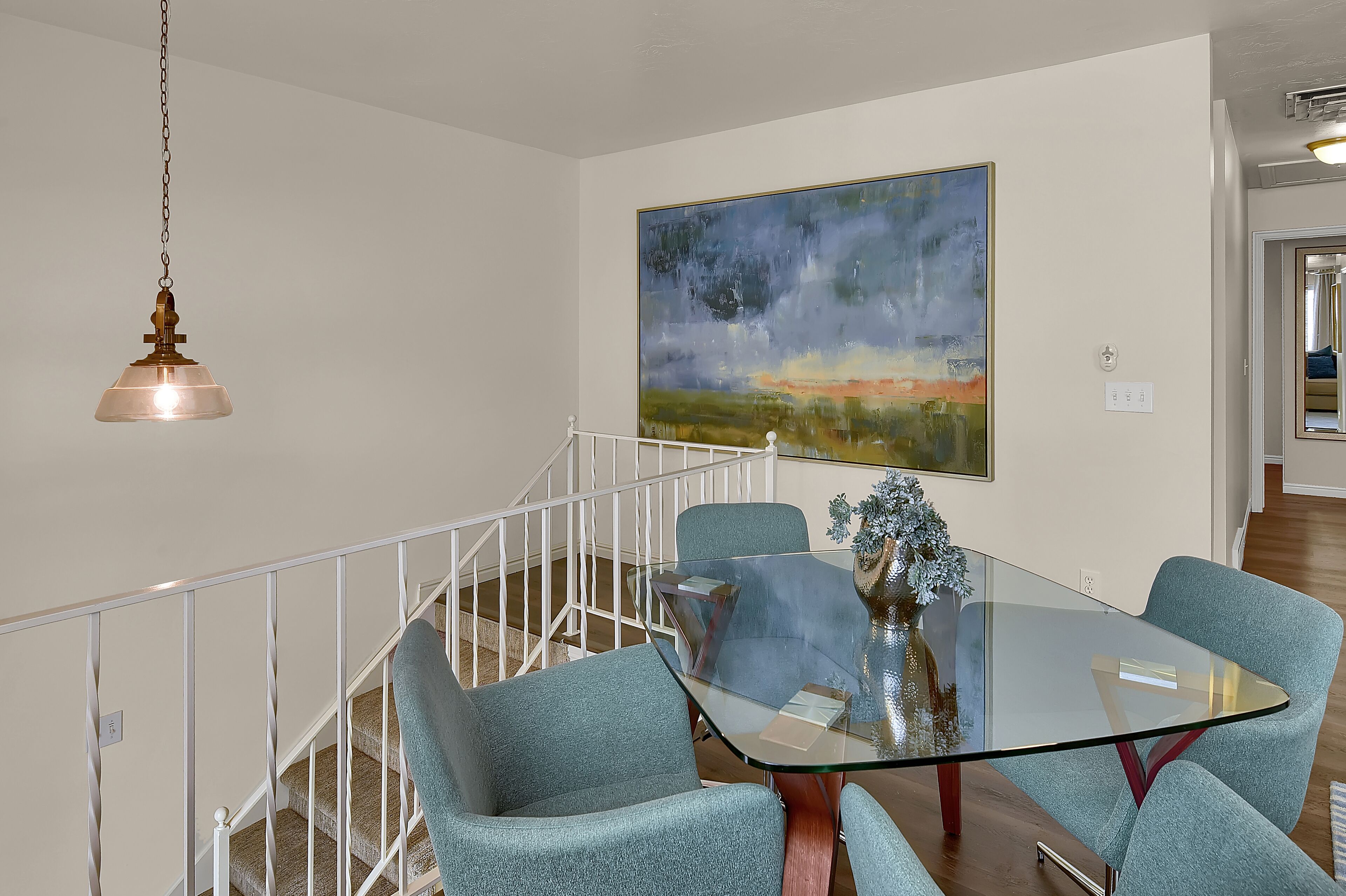 A dining area with a glass table and four blue chairs, a painting on the wall, and a staircase with a railing.