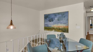 Un comedor con una mesa de vidrio y cuatro sillas azules, un cuadro en la pared y una escalera con barandilla.