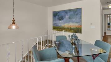 A dining area with a glass table and four blue chairs, a painting on the wall, and a staircase with a railing.