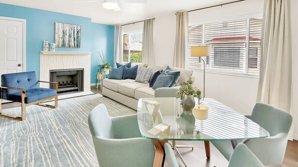 Una sala de estar moderna con una pared azul decorativa, una chimenea, un sofá con muchos cojines, una mesa de comedor de vidrio y un ventilador de techo.