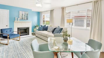 A modern living room with a blue accent wall, a fireplace, a sofa with multiple cushions, a glass dining table, and a ceiling fan.