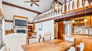 A spacious living area with a wooden dining table, cushioned seating, a fireplace, and a kitchen with wooden cabinets and pendant lighting. - Lakefront Cottage w Fireplace, Private Lake Access, Dock, Big Yard (Albrightsville)