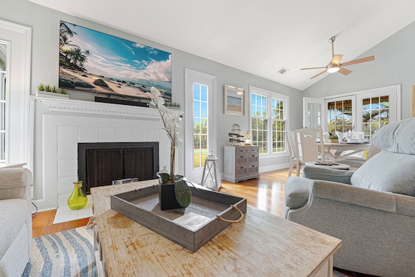 A spacious living room with a fireplace, a large painting, and a ceiling fan. - Shipyard Golf View Condo | Porch, Fireplace & Beach Access (Hilton Head Island)
