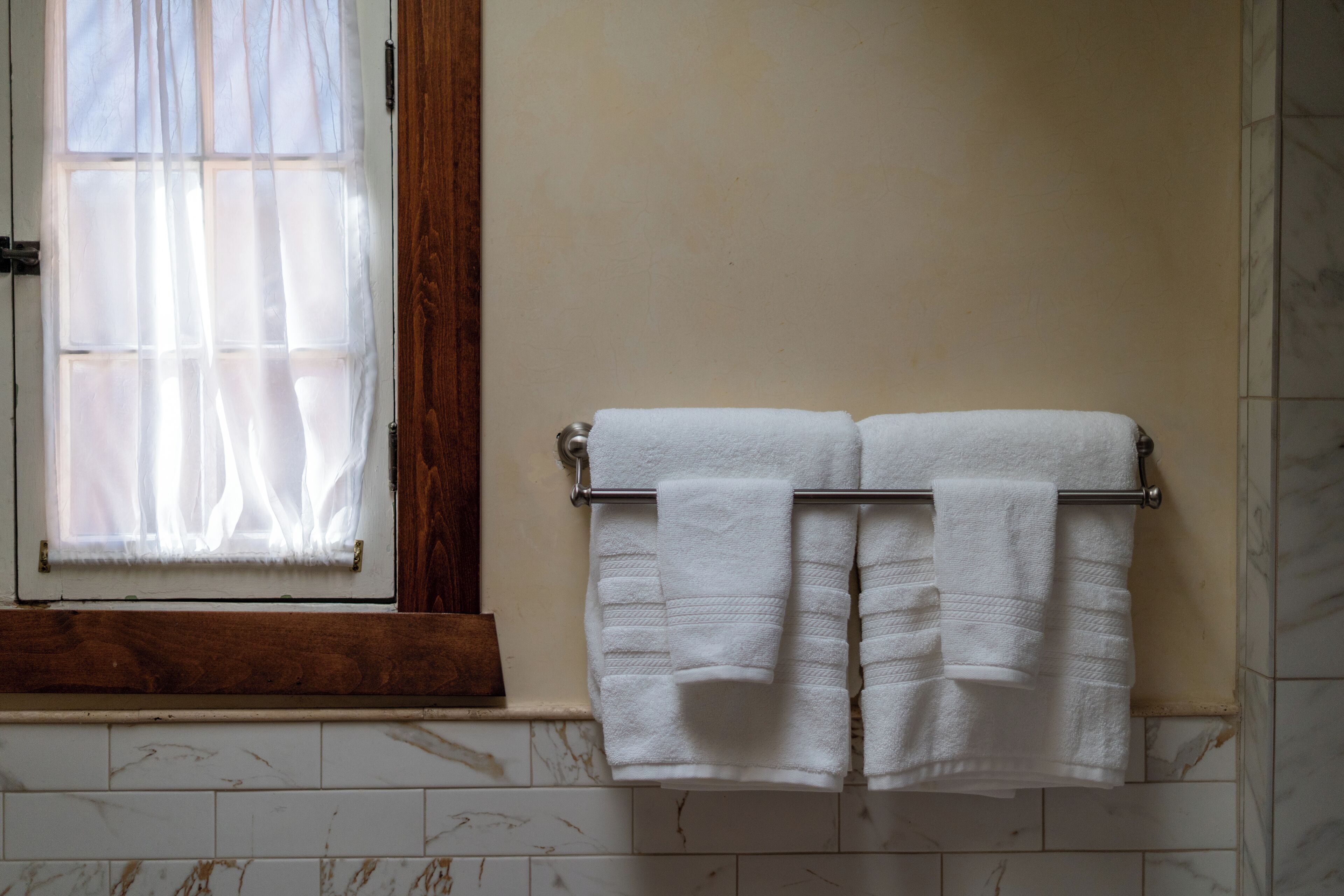 Un bagno con una finestra con stipite in legno, un portascopevoli con due asciugamani bianchi e un pavimento piastrellato.