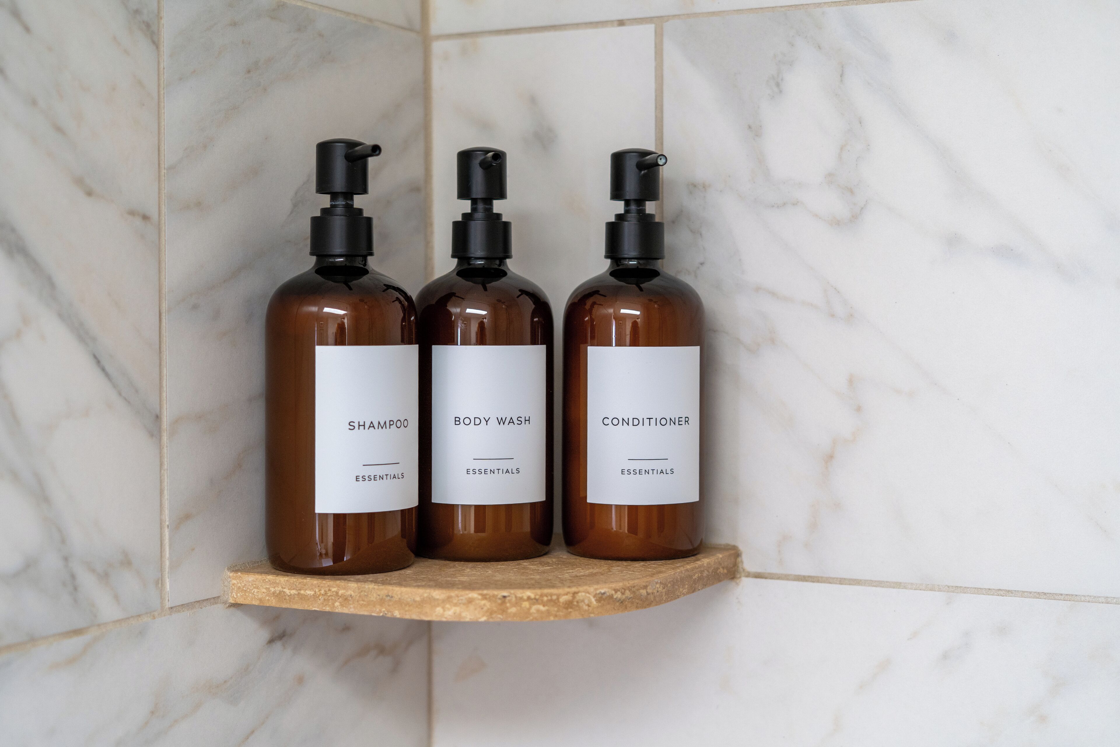 Three brown bottles of shampoo, body wash, and conditioner on a wooden shelf against a white marble wall.