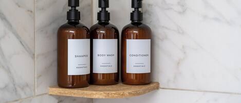 Three brown bottles of shampoo, body wash, and conditioner on a wooden shelf against a white marble wall.