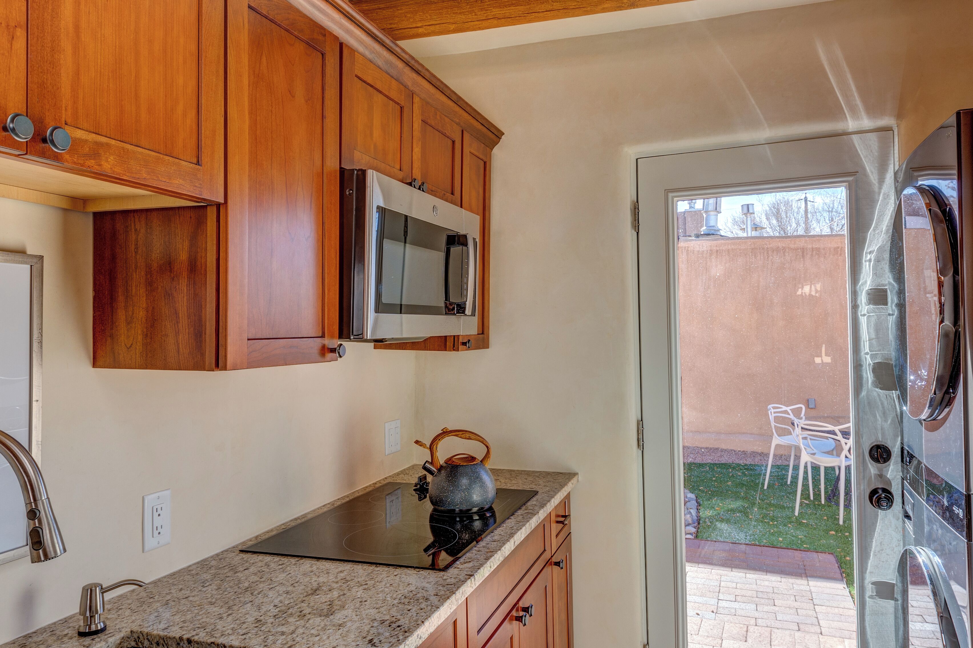 A kitchen with wooden cabinets, a microwave, a kettle, and a view of the backyard through a glass door.