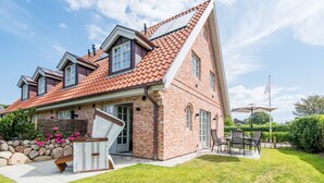 A two-story brick house with a sloped roof, a stone wall, and a patio with outdoor furniture. - Leev Marie, Germany (Sylt)