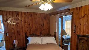 A bedroom with a wooden paneled wall, a bed with a floral bedspread, a ceiling fan, and a nightstand with a lamp.