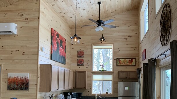 A wooden cabin interior with a kitchen, bar stools, a couch, and a ceiling fan.