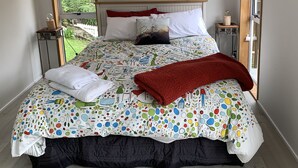 Un lit bien fait, recouvert d’une courtepointe à pois colorés, d’un plaid rouge et d’un coussin blanc orné de motifs floraux. Deux tables de chevet avec des lampes se trouvent de part et d’autre du lit, et une fenêtre offre une vue sur la verdure à l’extérieur.