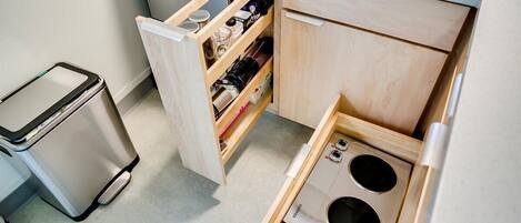 Een kleine keuken met een inbouwfornuis, een afvalbak en een houten kast met planken.