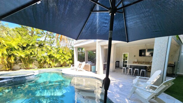 A swimming pool with a tiled edge, surrounded by a covered patio area with white lounge chairs and a bar.